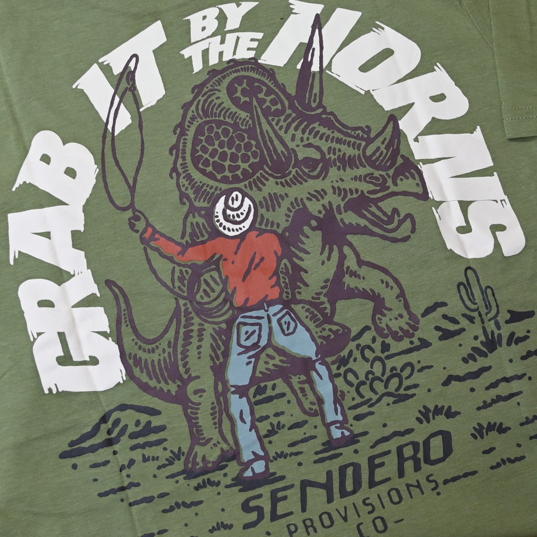 View of Sendero sage green Triceratops T-Shirt with cowboy roping a dinosaur graphic